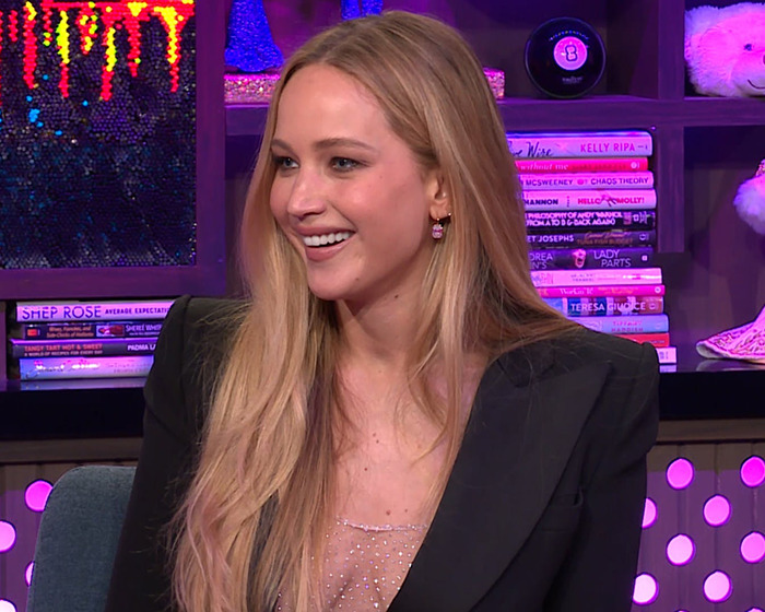 Smiling woman with long blonde hair on a talk show set, elegant attire, books and decorations in the background.