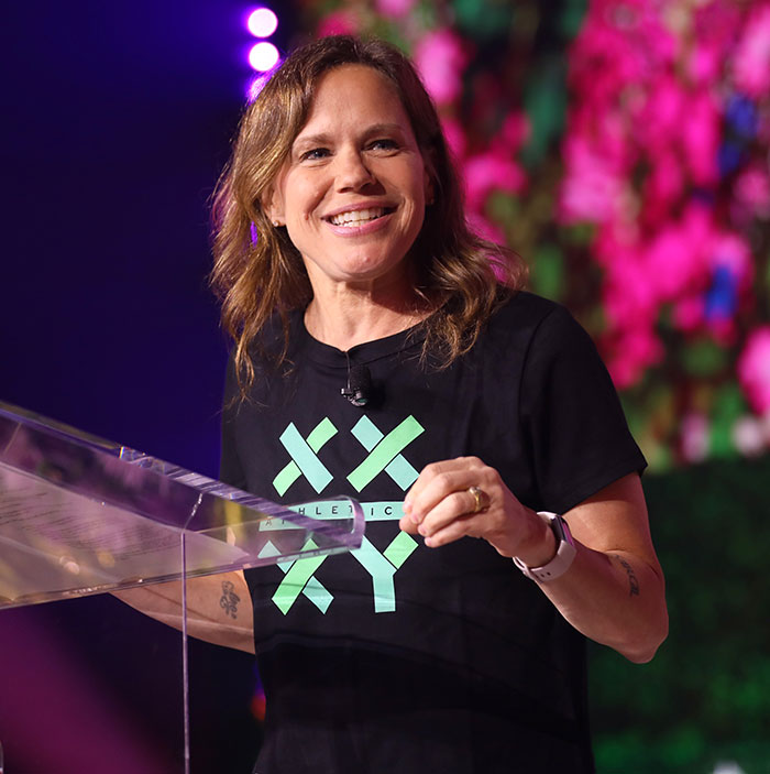Smiling individual in a black T-shirt speaking at a podium with colorful lights behind.