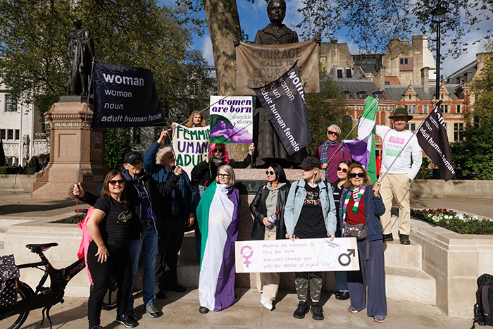 Protesters with signs on trans women and legal status outside a court, standing near a statue.