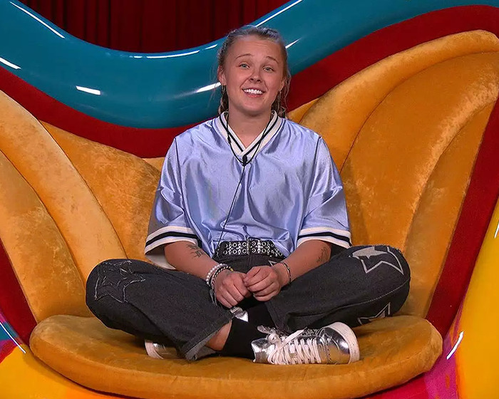 JoJo Siwa on a colorful couch, wearing a blue shirt and dark jeans, in a candid moment amid Big Brother discussions. JoJo Siwa on a colorful couch, wearing a blue shirt and dark jeans, in a candid moment amid Big Brother discussions.