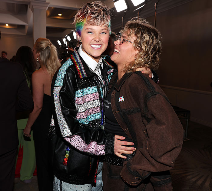 JoJo Siwa smiling at an event, wearing a colorful jacket, with a friend in a lively atmosphere.