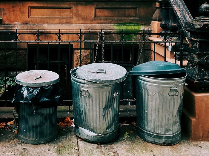 Three metal trash cans on a sidewalk, one overflowing, representing disgusting urban sights.