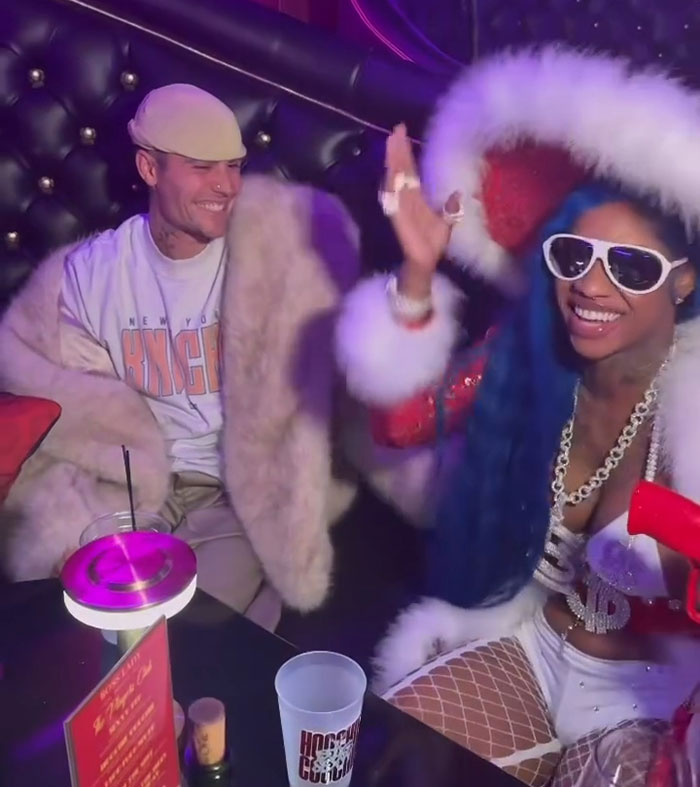 Celebrity smiling beside a female rapper in vibrant attire at a lively club setting.