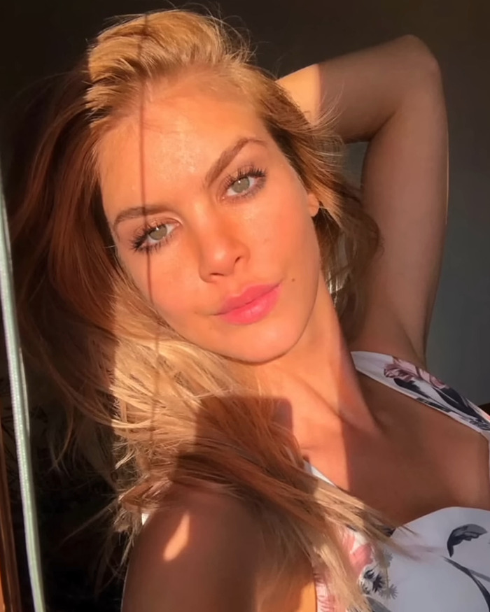 Blonde woman posing in sunlight with arm behind head, wearing a patterned top.