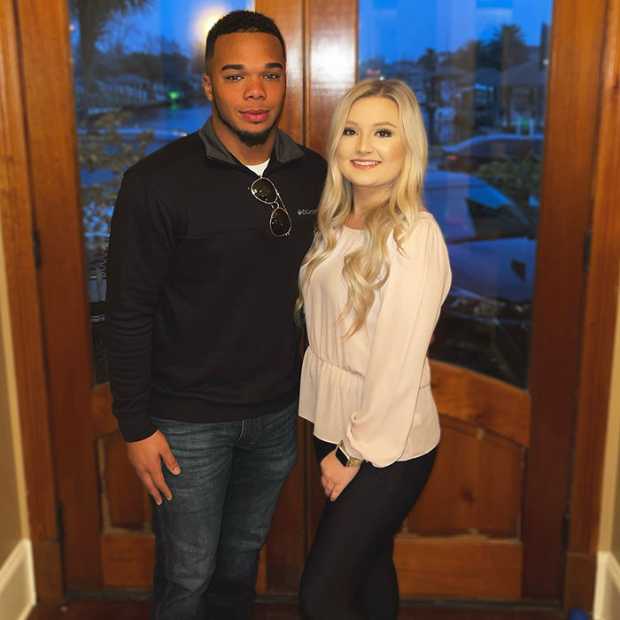 A couple standing together indoors, the man in a black jacket and the woman in a white blouse, smiling at the camera.