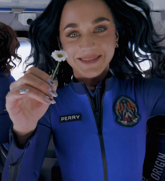 Woman in blue suit holding a small flower, smiling, with dark hair; patch visible on outfit. Woman in blue suit holding a small flower, smiling, with dark hair; patch visible on outfit.