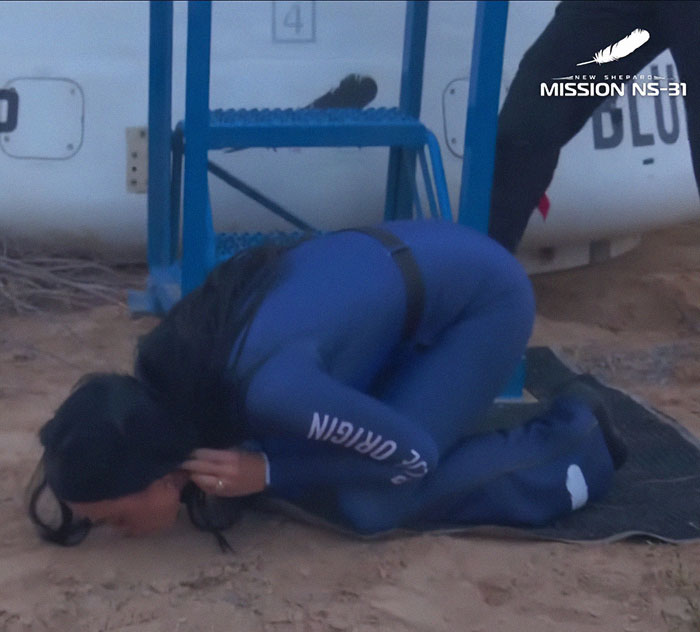 Person in a blue jumpsuit crouching on the ground, with "New Shepard" visible on the sleeve, near a space mission setup.