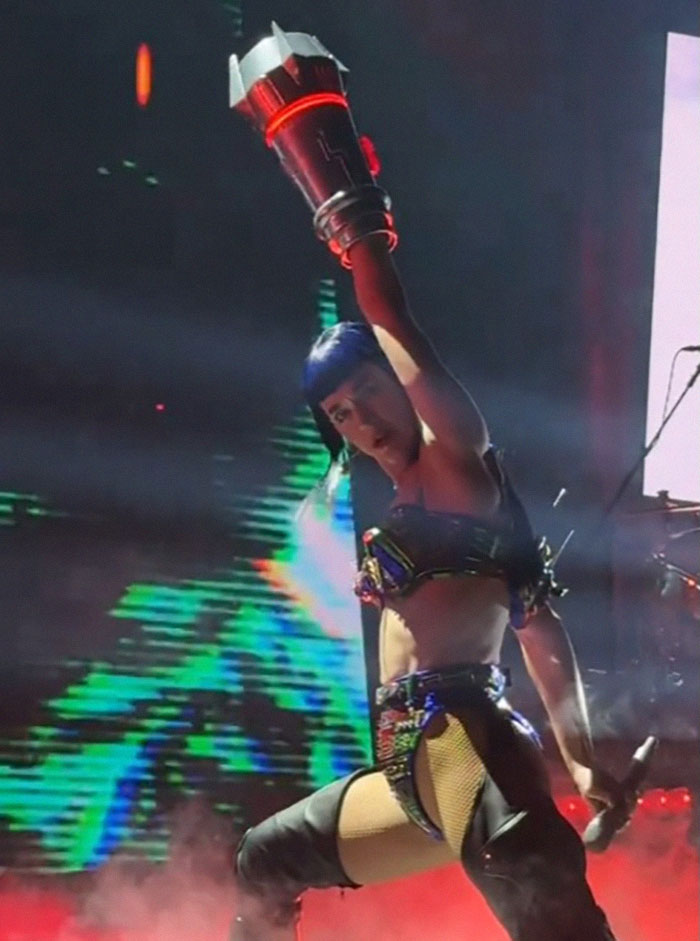 Singer performing on stage in futuristic attire, holding a microphone, with colorful lights in the background.