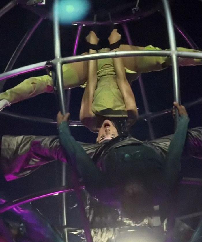 Singer performs a daring move upside down in a metal contraption during a tour, showcasing energetic dance choreography.