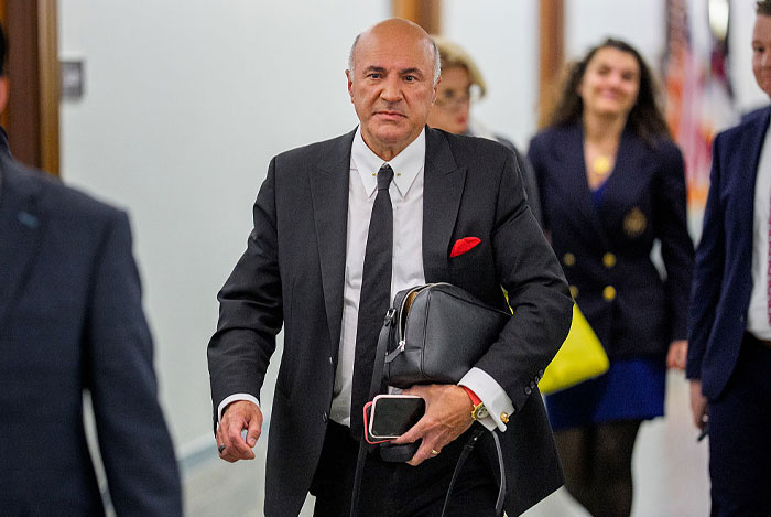 Kevin O&rsquo;Leary in a black suit carrying a bag and phone, walking confidently, highlighting his net worth and business success.