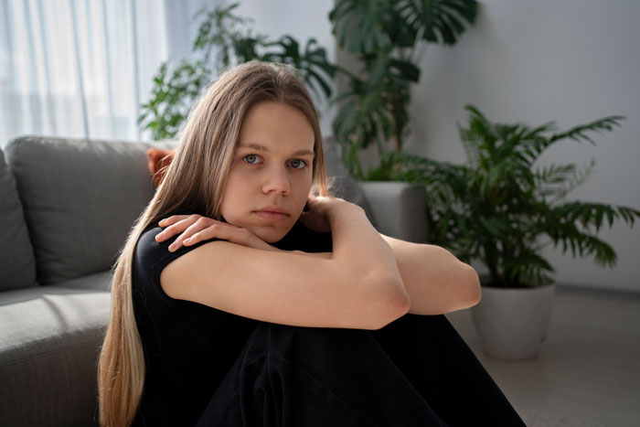 Teen sitting on a couch looking concerned in a living room, surrounded by green plants.