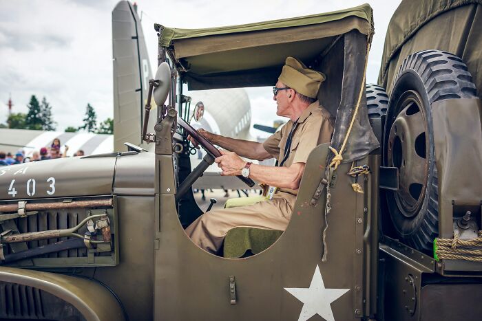A man in vintage military uniform drives an old army truck at a historical event, showcasing World War II machinery.