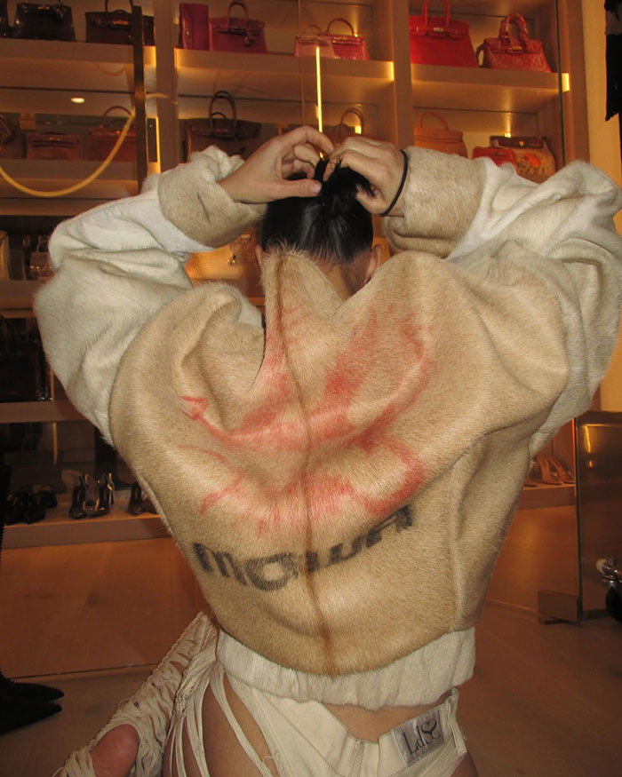 Woman in a furry coat adjusts her hair; shelves with handbags in the background.
