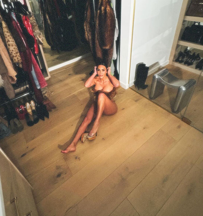Person posing in a closet setting, sitting on the floor, with fashionable attire and heels, emphasizing glamour.
