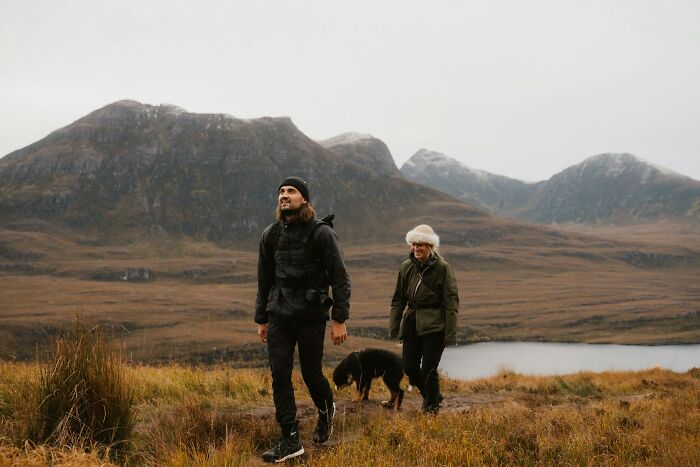 Two people hiking with a dog on a scenic island trail, surrounded by mountains and fields.