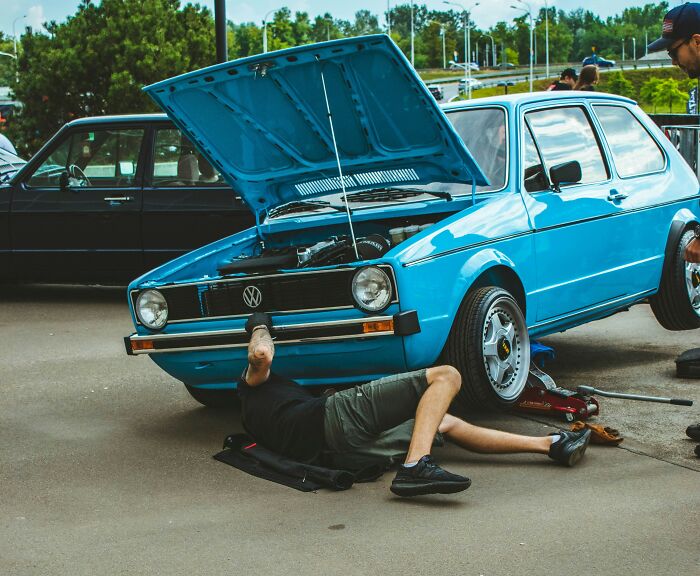 Man working under a blue car at an outdoor event, highlighting unique American habits.
