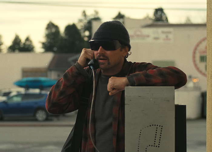 Man in casual attire, sunglasses, and beanie uses a payphone, showcasing a new look in an urban setting.