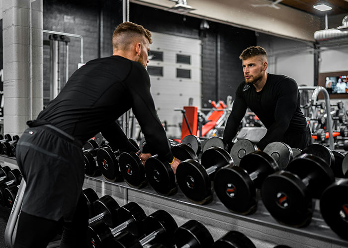Man in a gym, staring at his reflection in the mirror, pondering lifestyle choices, surrounded by dumbbells.