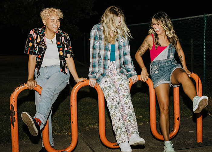 Three friends sitting on orange bars outdoors at night, laughing and showcasing carefree lifestyle choices.