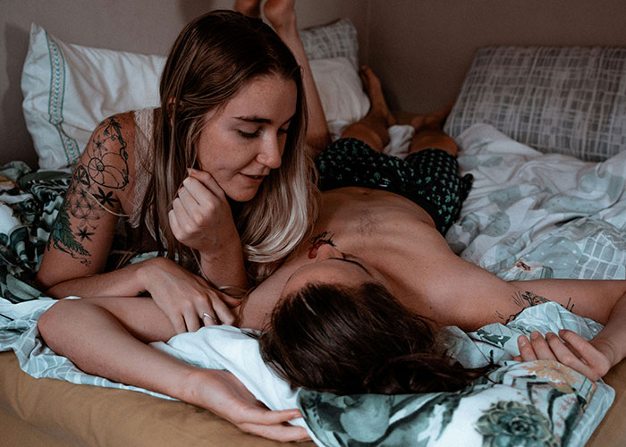 Couple lounging in bed, embracing lifestyle choices, with tattoos visible, suggesting potential relationship red flag.