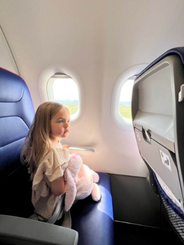 Child on airplane seat with plush toy, embodying youthful comedic charm in a travel setting.