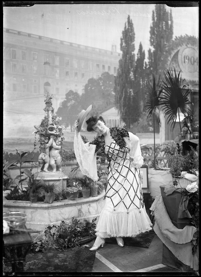 Woman in vintage dress tipping hat, posing in garden with plants and classical statue, rare photograph from glass negatives.