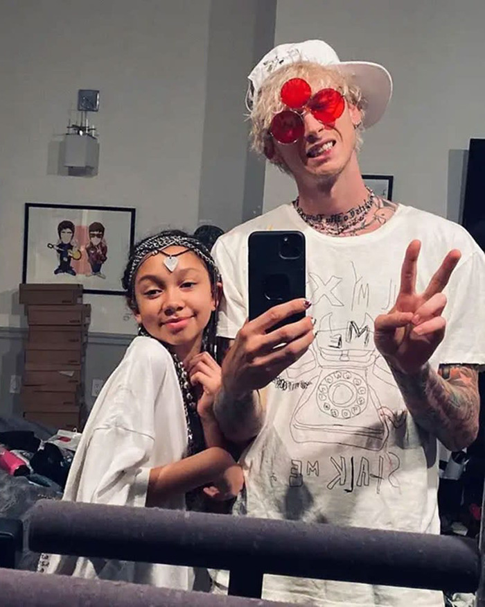 Machine Gun Kelly taking a selfie, wearing red glasses and a white hat, making a peace sign with a young girl.