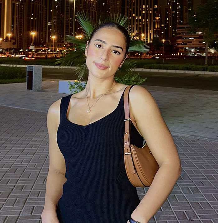 Woman in a black dress smiling at night with city lights in background, holding a brown purse.