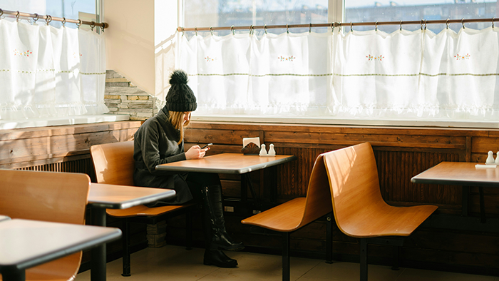 Woman sitting alone in a restaurant booth, wearing a winter hat and coat, looking at her phone by the window. Woman sitting alone in a restaurant booth, wearing a winter hat and coat, looking at her phone by the window.
