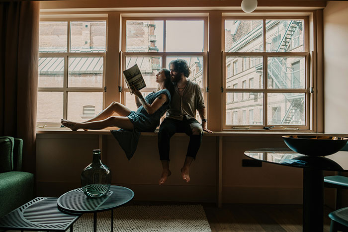 Couple sitting by large windows, with woman reading and man listening, embodying the concept of be present and connection.