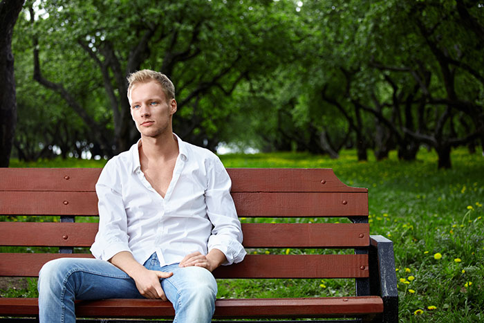Young man sitting on a park bench in a peaceful setting, wearing a white shirt and jeans, surrounded by lush greenery.