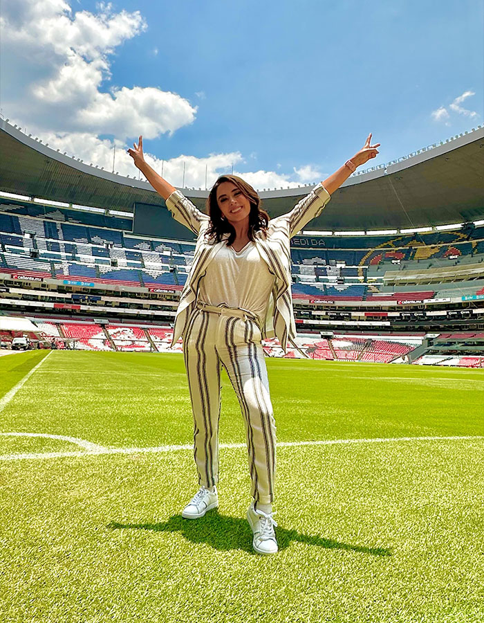 Female reporter stands on a soccer field, arms raised, smiling widely at the stadium.