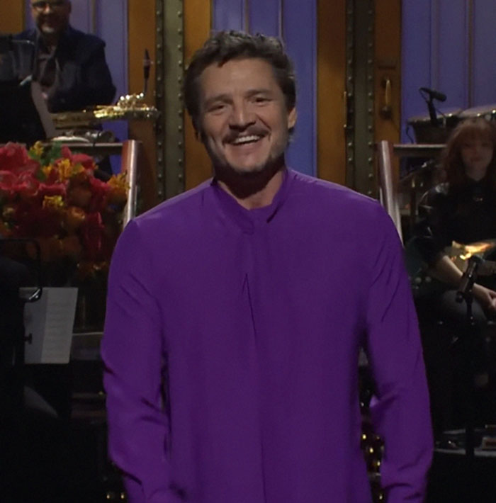 Man in a purple shirt smiling on stage during a live performance, band playing in the background. Celebrity encounter.