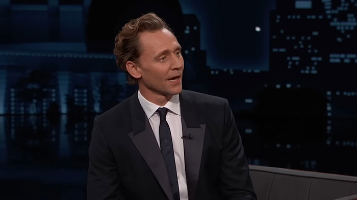 A celebrity in a dark suit speaking on a talk show set, with cityscape background.