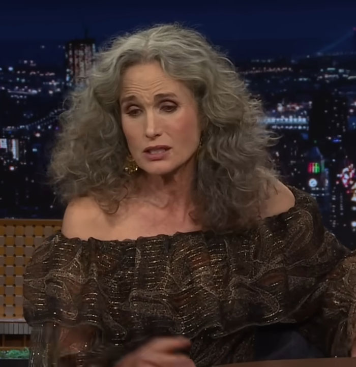 Woman with curly gray hair in off-shoulder top on a talk show, night cityscape in background, discussing celebrity encounters.