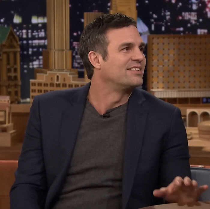 A man in a dark jacket, sitting and smiling during a television interview, with a cityscape backdrop.