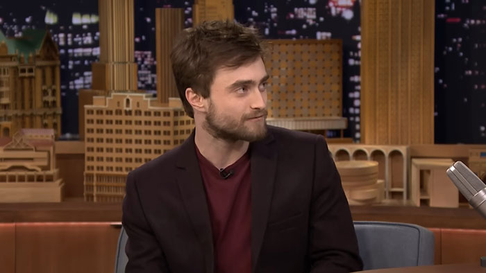 A celebrity encounter on a talk show, featuring a guest in a dark jacket and red shirt, set against a cityscape backdrop.