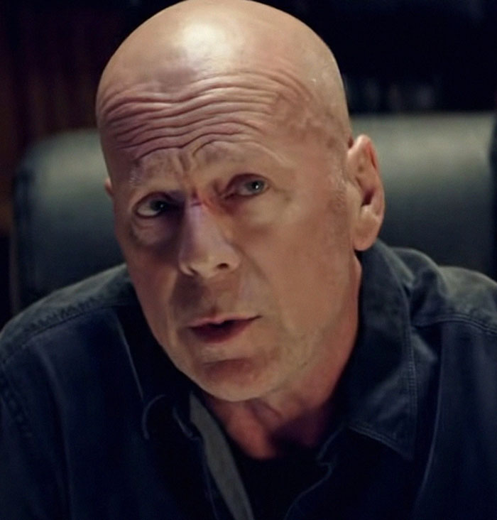 Bald man in a dark shirt, sitting indoors, looking thoughtfully.