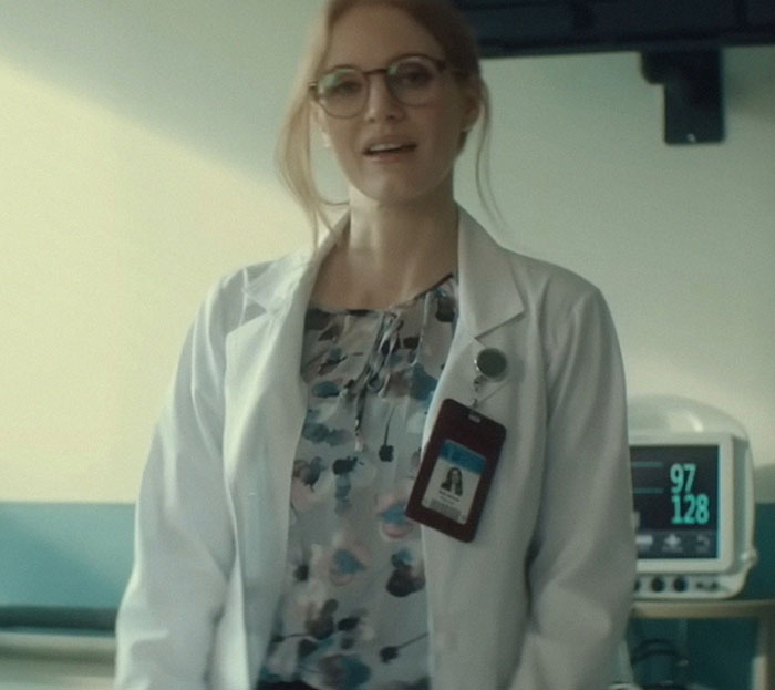 A woman in a lab coat and glasses stands in a room, wearing a floral blouse and ID badge.