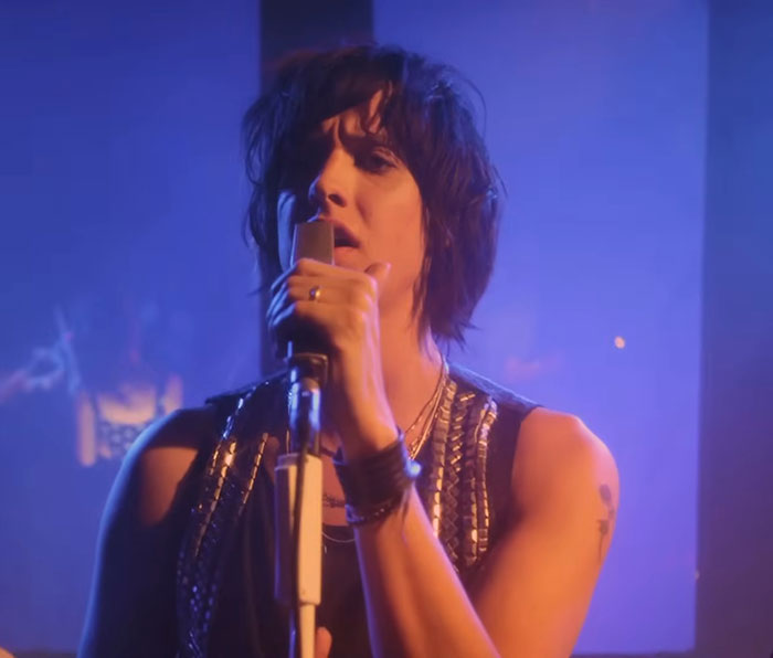 Singer performing on stage with a microphone, wearing a sleeveless top, in a vibrant blue-lit setting.