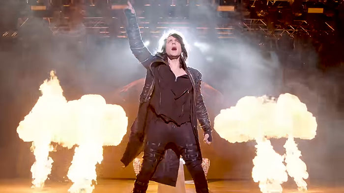 Celebrity performing on stage with fire effects, wearing a black outfit and raising one hand.