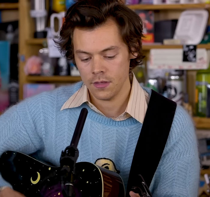 Celebrity playing acoustic guitar, wearing a light blue sweater, in a cozy setting.