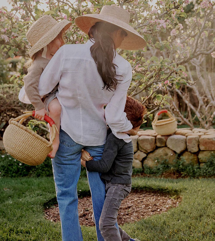Meghan Markle with Princess Lilibet and Prince Archie in garden, wearing hats and casual attire, enjoying a sunny day.