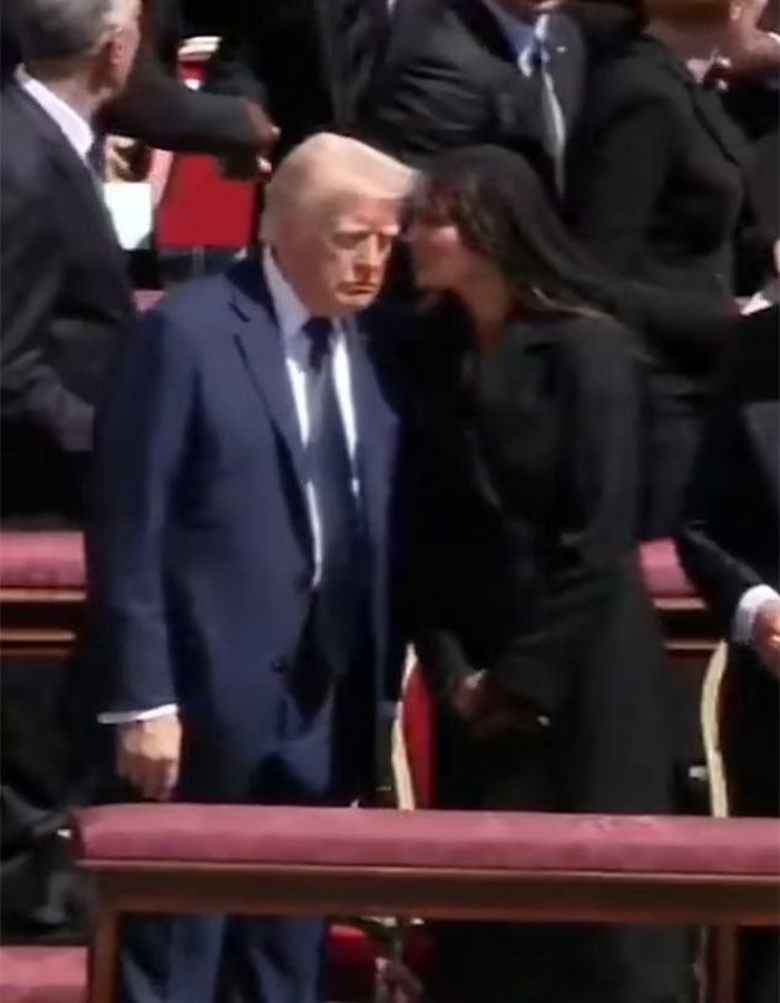 A woman whispers to a man in a suit at a formal event, with their heads close together, in a crowded setting.