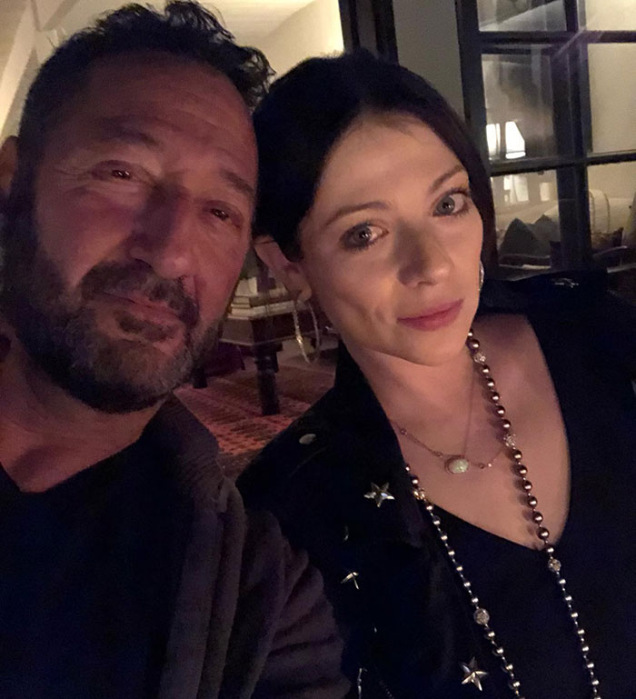 A woman in a black top with a beaded necklace, seated next to a man, in a dimly lit room. A woman in a black top with a beaded necklace, seated next to a man, in a dimly lit room.