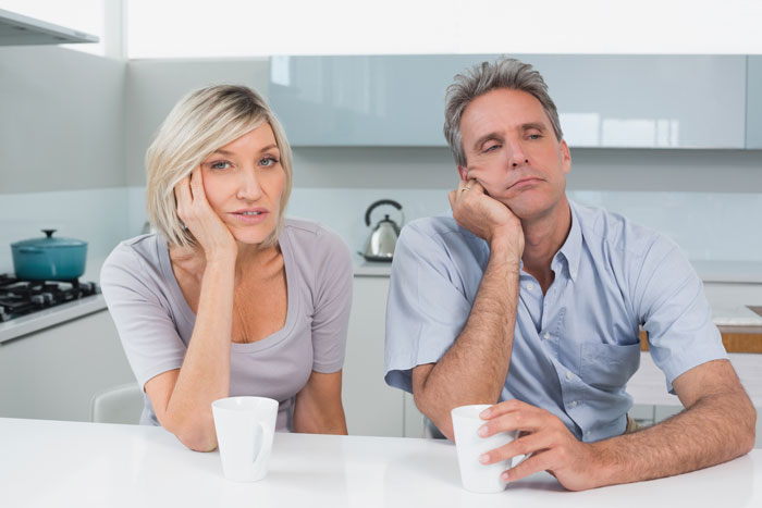 Man and woman sitting at a kitchen table with cups, appearing bored and pensive.