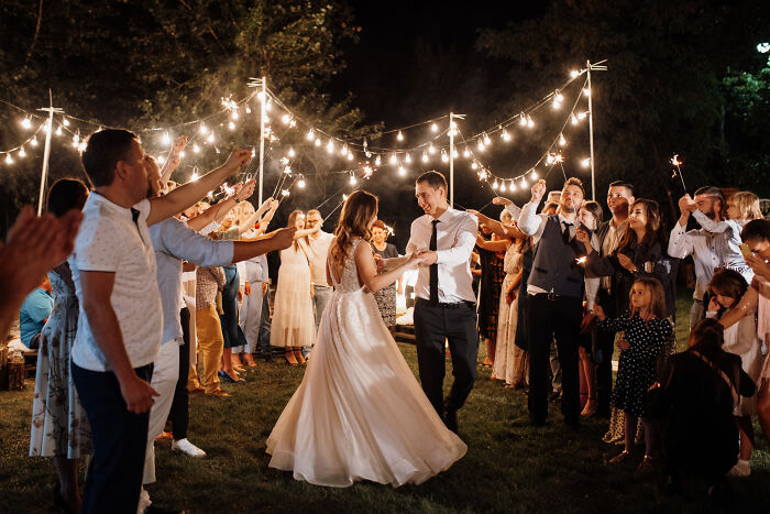 Couple dancing under string lights at an outdoor wedding reception surrounded by guests holding sparklers.