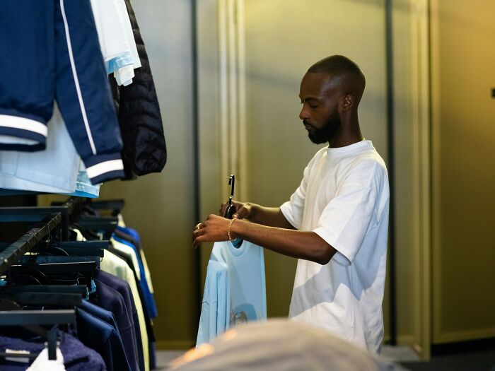 Elder millennial in a clothing store, selecting a blue shirt from a rack, reminiscent of 2008 recession struggles.