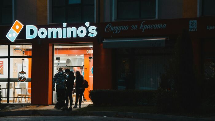 People entering a Domino's pizza store at night, the sign brightly lit against a dark backdrop.