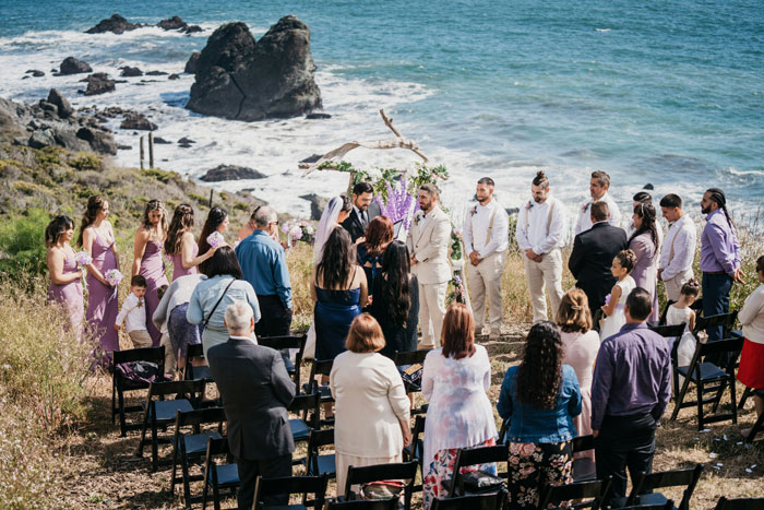 Wedding ceremony by the ocean with guests and a couple exchanging vows, highlighting parental gift demand issues.